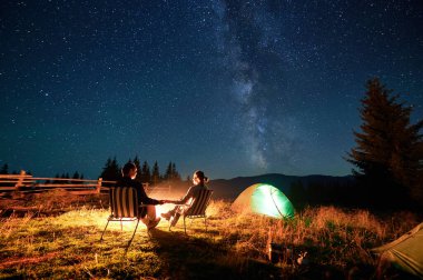 İki turist, kamp ateşi altında koltuklarda dinleniyor. Yanan çadırları, nefes kesen yıldızlı gökyüzünün altında. Samanyolu, dağ yabanda büyülü ve huzurlu bir atmosfer yaratarak yukarılara uzanır..