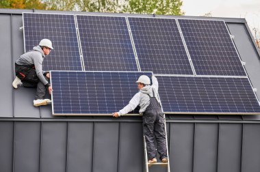 Çatıya fotovoltaik güneş modülü istasyonu inşa eden atçılar. Miğferli elektrikçiler güneş paneli sistemi kuruyor. Alternatif ve yenilenebilir enerji kavramı.
