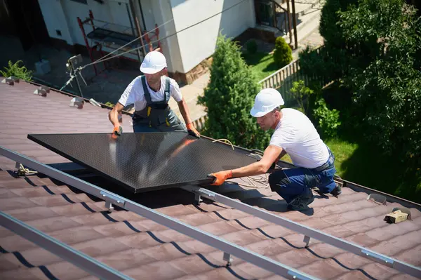 İşçiler evin çatısına güneş paneli inşa ediyorlar. Dışarıda fotovoltaik güneş modülü kuran kask takan iki adam. Alternatif, yeşil ve yenilenebilir enerji üretimi kavramı.