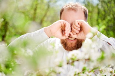 Baharda baharda çiçek açan bahçede mevsimsel alerji yüzünden acı çeken bir adam. Sakallı adamın hapşırması ve çiçek açan ağacın önünde kaşınması. Bahar alerjisi konsepti.