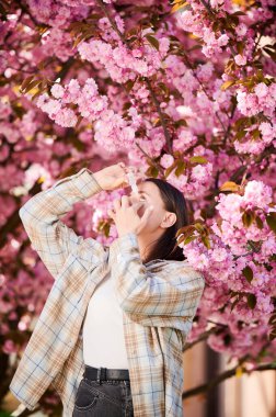 Tıbbi göz damlası kullanan alerjik bir kadın baharda çiçek açan bahçede mevsimsel alerjiden muzdarip. Açık havada çiçek açan ağacın önünde gözleri tedavi eden genç bir kadın. Bahar alerjisi konsepti.