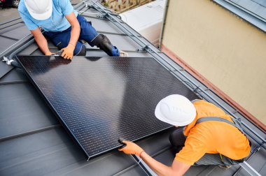 İşçiler evin çatısına fotovoltaik güneş paneli inşa ediyorlar. Miğferli teknisyenler ve eldivenli adamlar dışarıda büyücülerin yardımıyla güneş modülü kuruyorlar. Yenilenebilir enerji üretimi kavramı.