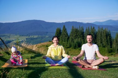 Genç bir aile sabahları doğa manzaralı dağlarda yoga yapıyor. Baba, anne ve kız yoga minderlerinde, arka planda dağları ve ormanı manzaralı açık mavi gökyüzünün altında.