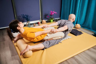Masör Tayland yogası masajı yapıyor. Geleneksel masaj tedavisi uygulayan kadın terapist, erkek hastaların vücudunu esnetiyor. Kel müşteri paspasın üzerinde uzanıyor, sakin atmosferde el masajının keyfini çıkarıyor..