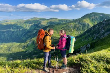 İki yürüyüşçü, turuncu sırt çantalı bir adam ve yeşil yastıklı pembe ceketli bir kadın, dağ yolunda karşı karşıya duruyorlar. Arka planda parlak mavi gökyüzünün altındaki göz kamaştırıcı yeşil tepeler.