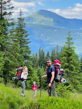 Üç kişilik bir aile, büyük sırt çantaları taşıyan iki yetişkin ve pembe ceketli ve şapkalı bir çocuk otlak yamaçta duruyor. Sersemletici dağ manzarası yemyeşil ağaçlar ve arka planda açık mavi gökyüzü.
