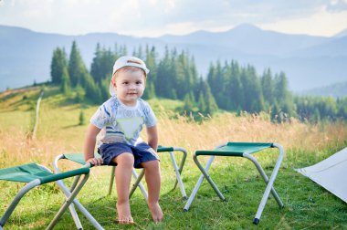 Genç çocuk çimenli alanda küçük katlanan taburede yalınayak oturuyor, yanında da iki boş tabure var. Beyaz şapkalı ve desenli gömlekli. Bereketli, yeşil tepeler ve berrak gökyüzünün altındaki dağlar.