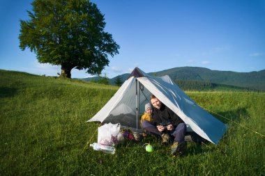 Mutlu baba ve çocuğun portresi arka planda manzaralı dağlarla otlak yamaçtaki beyaz çadırda oturuyor. Büyük ağaç ve kamp malzemeleri açık mavi gökyüzünün altında..