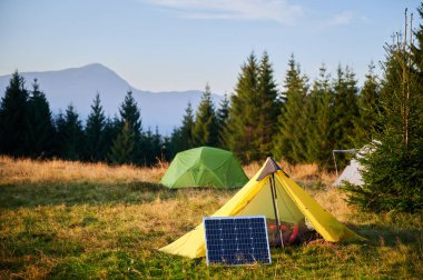 Arka planda çadırları olan çimenli alandaki güneş paneli, akşamları sık yeşil ağaçlarla çevrili çevre dostu kamp yerlerini ön plana çıkarıyor. Açık hava faaliyetlerinde yenilenebilir enerji entegrasyonu.