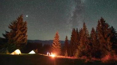 Dağlarda gece kampı. Ormanlı dağ manzarasının ortasında iki çadır parlak, yıldızlarla dolu gökyüzünün altında kamp ateşinin yanında sıcacık parlıyor. Samanyolu 'nun altındaki kamp alanında dinlenen turistlerin siluetleri.