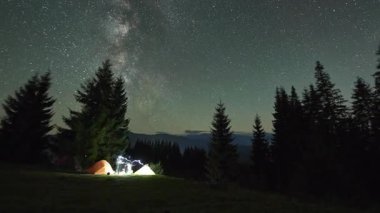 Dağlarda gece kampı. Ormanlı dağ manzarasının ortasında iki çadır parlak, yıldızlarla dolu gökyüzünün altında kamp ateşinin yanında sıcacık parlıyor. Samanyolu 'nun altındaki kamp alanında dinlenen turistlerin siluetleri.