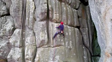 Erkek kaya tırmanıcısının engebeli kireçtaşı kayalıklara tırmanırken güvenlik için koşum takımı ve ip ile hava görüntüsü. Sporcu Ukrayna 'nın Karpatya dağlarındaki Dobvush Kayaları' nda dikey büyük kaya parçasına tırmanıyor.