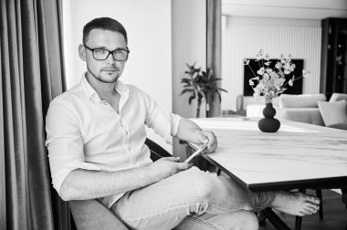 Akıllı telefonlu bir adamın portresi mutfaktaki beyaz mermer tezgahta oturuyor, gözlük takıyor. Dairesinde dinlenen yakışıklı bir adam. Tarz sahibi bir adam kameraya bakıyor. Siyah ve beyaz