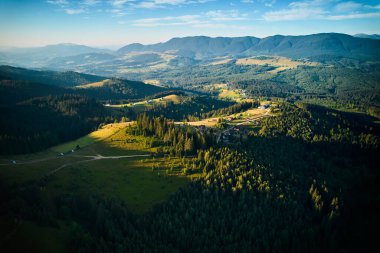 Yuvarlanan tepeleri, yemyeşil ormanları ve dağınık evleri olan gökyüzü manzaralı bir vadi. Güneş ışığı manzarayı ılık bir ışıltıyla yıkıyor, canlı yeşillikleri renklendiriyor ve sakin bir atmosfer yaratıyor..