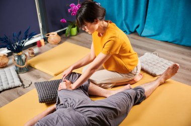Masör Tayland yogası masajı yapıyor. Geleneksel masaj tedavisi uygulayan kadın terapist, erkek hastaların vücudunu esnetiyor. Müşteri paspasın üzerinde uzanıyor, sakin atmosferde el masajının keyfini çıkarıyor..