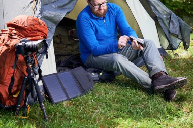 Sakallı adam otlak bölgedeki turist çadırının girişinde oturuyor ve elektrik bankasına taşınabilir güneş paneli yüklüyor. Yakınlarda, üç ayaklı kamera ve turuncu sırt çantası fotoğrafçılığa olan ilgisini gösteriyor..