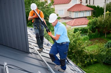 İşçiler evin çatısına güneş paneli inşa ediyorlar. Dışarıda fotovoltaik güneş modülü taşıyan kasklı iki adam var. Alternatif, yeşil ve yenilenebilir enerji üretimi kavramı.