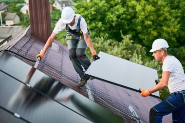 İşçiler evin çatısına güneş paneli inşa ediyorlar. Dışarıda fotovoltaik güneş modülü taşıyan kasklı iki adam var. Alternatif, yeşil ve yenilenebilir enerji üretimi kavramı.
