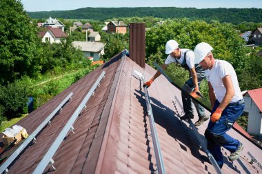 İşçiler evin çatısına güneş paneli inşa ediyorlar. Dışarıda fotovoltaik güneş modülü taşıyan kasklı iki adam var. Alternatif, yeşil ve yenilenebilir enerji üretimi kavramı.