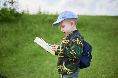 Genç bir çocuk çimenlik alanda yürüyüş yapıyor, dikkatle harita ve açık havada pusula çalışıyor. Omuzlarında sırt çantasıyla, turist dağlardaki rotasını belirlemeye veya yönlendirmeye odaklanmış durumda..
