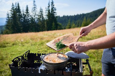 Kapalı ellere odun kesme tahtasından kesilmiş yeşillikleri açık hava sobasında kaynayan tencereye ekleyin. Sahne çimenli çayırlarda, arka planda uzun çam ağaçları, berrak mavi gökyüzünün altında.