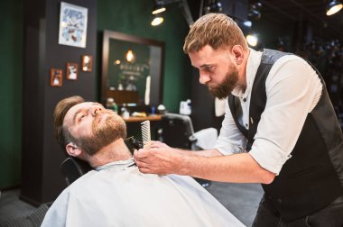 Profesyonel berber müşterinin sakalını kesiyor. Erkek kuaför, kırpıcının ince sakallı müşterisine servis yapıyor. Adamın saçını kestirirken çekilmiş bir fotoğrafı..