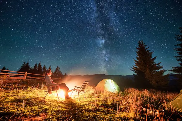 Kamp ateşinde dinlenen turist, sıcak bir ışıltıyla güneşleniyor, nefes kesici bir çadırın altında, yıldızlarla dolu bir gökyüzü. Samanyolu uzanıyor, huzurlu ve büyülü dağ manzarası yaratıyor..