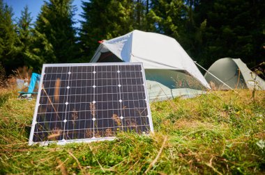 Arka planda çadırları olan çimenli alandaki güneş paneli, sabah sık yeşil ağaçlarla çevrili çevre dostu kamp düzeneğini ön plana çıkarıyor. Açık hava faaliyetlerinde yenilenebilir enerji entegrasyonu.