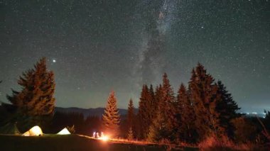 Dağlarda gece kampı. Ormanlı dağ manzarasının ortasında iki çadır parlak, yıldızlarla dolu gökyüzünün altında kamp ateşinin yanında sıcacık parlıyor. Samanyolu 'nun altındaki kamp alanında dinlenen turistlerin siluetleri.