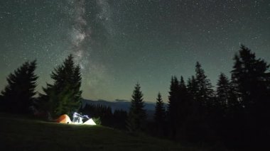 Dağlarda gece kampı. Ormanlı dağ manzarasının ortasında iki çadır parlak, yıldızlarla dolu gökyüzünün altında kamp ateşinin yanında sıcacık parlıyor. Samanyolu 'nun altındaki kamp alanında dinlenen turistlerin siluetleri.