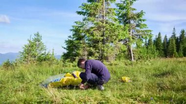 Zaman aşımı. Yürüyüşçü, çim yamacında çadır kurmaya çalışıyor. Çam ağaçları ve uzak dağlar parçalı bulutlu gökyüzünün altında.