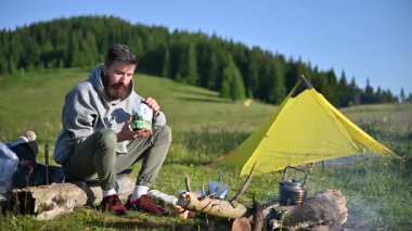 Yeşil kaşıkla yemek kesesini yiyen bir turist kamp ateşinin yanındaki kütüğe oturur. Yeşil yamaçta, yemyeşil ağaçlarla çevrili sarı çadır. Güzel doğal ortamda kamp yapmanın huzuru..