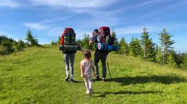 Küçük çocuk da dahil olmak üzere yürüyüşçülerin ailesinin arka manzarası güneşli çayırlarda yürüyor. Turistler kamp malzemeleriyle birlikte büyük sırt çantaları taşırken, kızı da onu yakından takip ediyor..