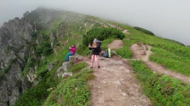 Hava manzaralı iki yürüyüşçü sisli dağ yolunda fotoğraf çekiyor, yürüyüş direkleri ile duruyor. Etrafı yemyeşil bitki örtüsü ve engebeli uçurumlarla çevrili kadın turistler sisli manzaranın güzelliğini yakalıyorlar..