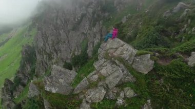 Yürüyüşçünün hava manzarası, dik ve sivri uçurumları olan sis kaplı vadiye tepeden bakan büyük bir kayanın üzerindedir. Aşağıdaki yemyeşil yamaçlar siste kısmen görülebilir, dingin ve mistik bir atmosfer yaratıyor..