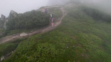 Üç yürüyüşçünün hava manzarası sisli dağ sırtı boyunca yüksek kaya oluşumlarıyla çevrili kuvvetli rüzgarla yürüyor. Kayalık arazi ve yemyeşil bitki örtüsü dingin bir atmosfer yaratır.