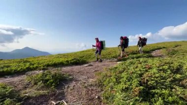 Üç yürüyüşçü parçalı bulutlu gökyüzünün altında yeşil dağ yolu boyunca yürürler. Yuvarlanan tepeler ve uzak tepeler nefes kesici bir zemin oluşturuyor. Yürüyüşçüler iyi donanımlı, temiz ve manzaralı yolculuğun tadını çıkarıyorlar..
