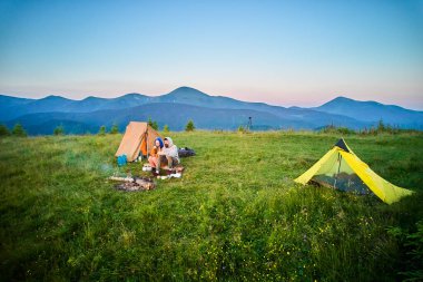Sabahları iki çadırı olan manzaralı dağ kampı. İki kampçı, bulutsuz gökyüzünün altında yuvarlanan dağların huzurlu manzarasının keyfini çıkarıyorlar. Resimli gündoğumu manzarası, huzurlu ve rahat bir açık hava deneyimi yaratıyor..