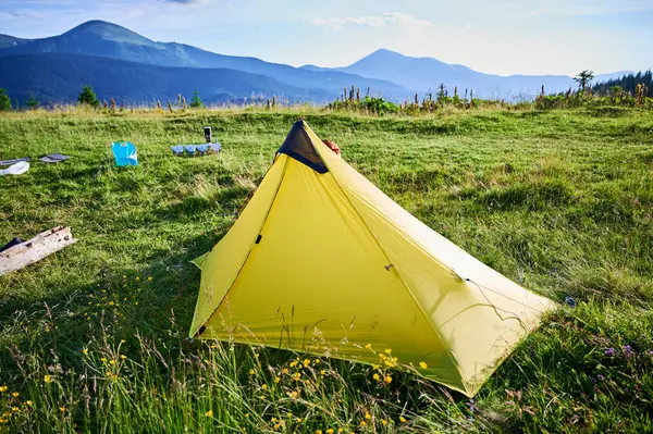 Çimenli yamaçta sarı turist çadırı. Kamp malzemeleri de yakınlarda. Sersemletici yeşil tepeler, berrak mavi gökyüzünün altındaki uzak dağlar, barışçıl açık hava maceraları için mükemmel bir yer yaratıyor..
