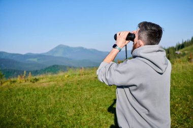 Adam çim tarlasında duruyor, uzak dağları gözlemlemek için dürbün kullanıyor, sol bileğinde gri kapüşonlu, akıllı bir şey var. Yuvarlanan yeşil tepeler ve berrak mavi gökyüzü, sakin bir manzara yaratıyor..