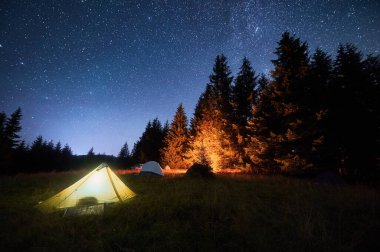 Çimenli, berrak, yıldızlı gökyüzünün altında parlayan kamp ateşiyle aydınlatılmış birkaç çadır. Dağlarda yüksek çam ağaçlarıyla çevrili. Doğada kamp yaparak geçirilen gecenin huzurlu güzelliği..