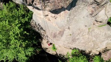 Erkek kaya tırmanıcısının engebeli kireçtaşı kayalıklara tırmanırken güvenlik için koşum takımı ve ip ile hava görüntüsü. Sporcu Ukrayna 'nın Karpatya dağlarındaki Dobvush Kayaları' nda dikey büyük kaya parçasına tırmanıyor.