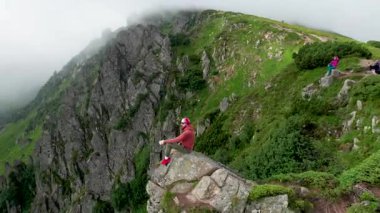 Yürüyüşçünün hava manzarası büyük bir kayanın üzerinde, aşağıdaki sisli, pürüzlü uçurumların ve yeşil vadinin nefes kesici manzarasının tadını çıkarıyor. Kırmızı kapüşonlu ve bandanalı, dingin, rüzgarlı manzaraya renk katıyor..