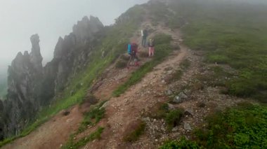 Üç yürüyüşçünün hava manzarası sisli dağ sırtı boyunca yüksek kaya oluşumlarıyla çevrili kuvvetli rüzgarla yürüyor. Kayalık arazi ve yemyeşil bitki örtüsü dingin bir atmosfer yaratır.