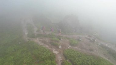 Üç yürüyüşçünün hava manzarası sisli dağ sırtı boyunca yüksek kaya oluşumlarıyla çevrili kuvvetli rüzgarla yürüyor. Kayalık arazi ve yemyeşil bitki örtüsü dingin bir atmosfer yaratır.