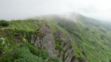 Yürüyüşçünün hava manzarası büyük bir kayanın üzerinde, aşağıdaki sisli, pürüzlü uçurumların ve yeşil vadinin nefes kesici manzarasının tadını çıkarıyor. Kırmızı kapüşonlu ve bandanalı, dingin, rüzgarlı manzaraya renk katıyor..