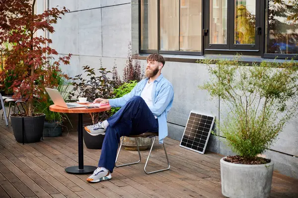 Mutlu sakallı adam yuvarlak tahta masada dizüstü bilgisayarı, defteri ve bir fincan kahveyle çalışıyor. Adam kulaklık takıyor, çevresi yeşillik ve güneş paneliyle çevrili, modern, üretken çalışma alanını vurguluyor..