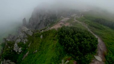Üç yürüyüşçünün hava manzarası sisli dağ sırtı boyunca yüksek kaya oluşumlarıyla çevrili kuvvetli rüzgarla yürüyor. Kayalık arazi ve yemyeşil bitki örtüsü dingin bir atmosfer yaratır.