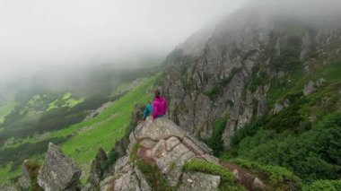 Yürüyüşçünün hava manzarası, dik ve sivri uçurumları olan sis kaplı vadiye tepeden bakan büyük bir kayanın üzerindedir. Aşağıdaki yemyeşil yamaçlar siste kısmen görülebilir, dingin ve mistik bir atmosfer yaratıyor..