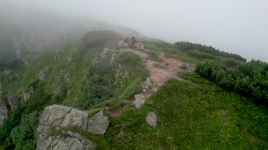 Üç yürüyüşçünün hava manzarası sisli dağ sırtı boyunca yüksek kaya oluşumlarıyla çevrili kuvvetli rüzgarla yürüyor. Kayalık arazi ve yemyeşil bitki örtüsü dingin bir atmosfer yaratır.